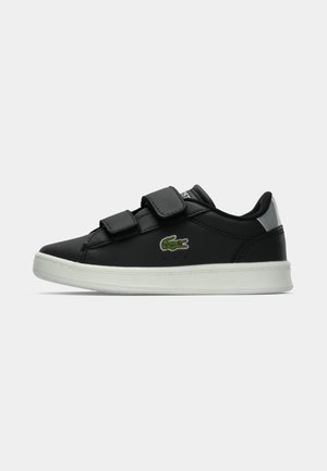 Zapato de cuero negro con dos correas de Velcro, suela de goma blanca y sutil logo de cocodrilo verde en el lateral. Presenta un diseño de puntera redondeada.
