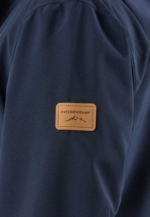 Marineblauwe stof met een met stiksel vastgenaaid bruin leren label met een bergafbeelding en de merknaam "Swedemount".