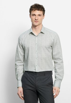 Uomo che indossa una camicia bianca a maniche lunghe con motivi, infilata in pantaloni grigio scuro, in piedi davanti a uno sfondo bianco semplice.