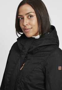 Veste noire isolante avec capuche, présentant un tissu texturé, une fermeture éclair à l'avant et une touche en bois près du col.