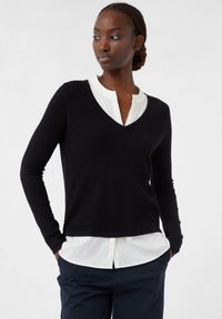 Frau mit geflochtenen Haaren, die einen schwarzen strukturierten V-Ausschnitt-Pullover über einem weißen Hemd trägt, die Hände in den Taschen von blauen Hosen, schaut nach links.