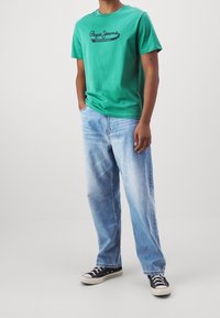 Camiseta de algodón verde con un logo negro en el frente, combinada con jeans rectos de corte azul claro y zapatillas negras.