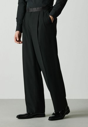 Person trägt schwarze plissierte weit geschnittene Hose, schwarzen Gürtel, schwarzes Hemd und glänzende schwarze Lederschuhe, steht mit einer Hand in der Tasche.