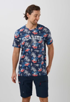 Lachende man die een blauw tropisch bloemen T-shirt met de tekst "SEASIDE" en marineblauwe shorts draagt tegen een effen lichte achtergrond.