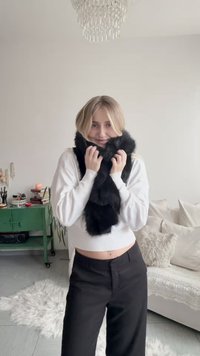Black faux fur scarf wrapped around the neck, worn with a white ribbed sweater and black high-waisted pants. Bright interior lighting.