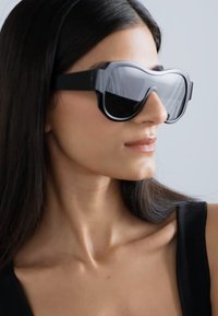 Occhiali da sole neri oversize con un design elegante, lenti ampie e un bordo bianco a contrasto. La montatura è liscia con una forma moderna e angolare.
