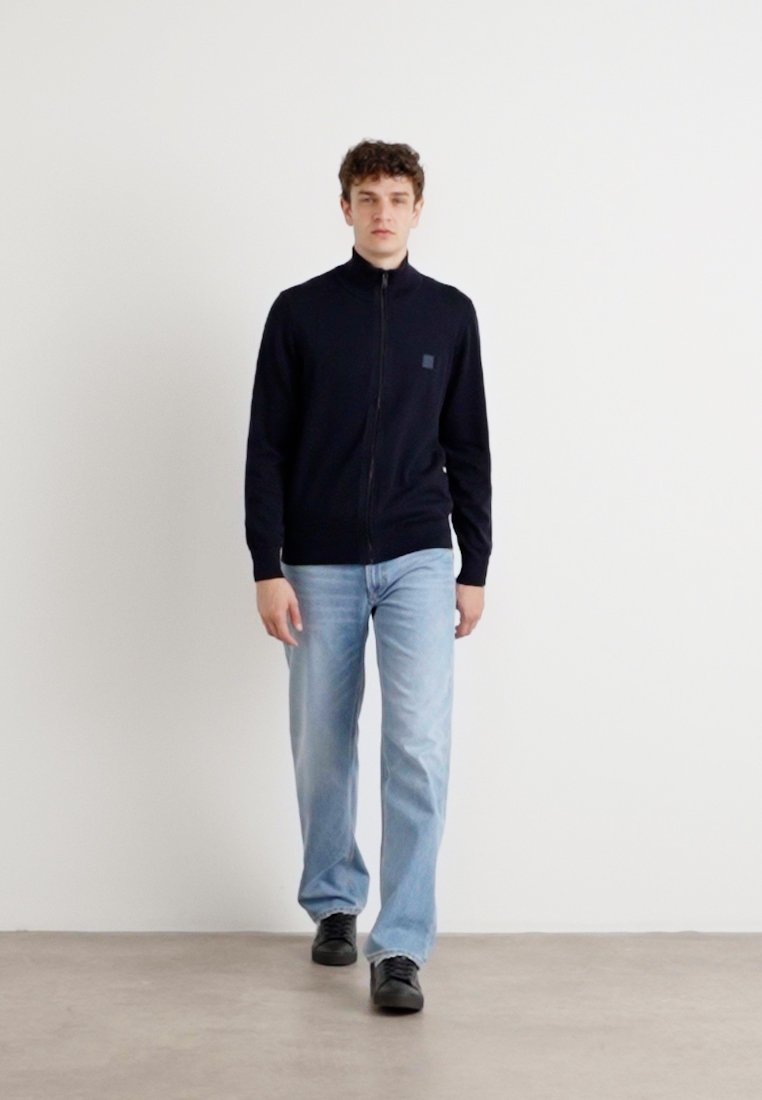 Sudadera azul marino con cremallera y cuello alto, puños acanalados y un pequeño parche con logo; combinada con vaqueros de mezclilla claros y zapatos negros.