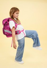 Rosa Rucksack mit grauen Akzenten, getragen von einem Kind. Das Kind trägt ein weißes T-Shirt und eine hellblaue Jeans mit Smileykopf-Grafiken. Der Hintergrund ist gelb.