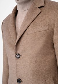 Bruine wollen blazer met een zachte textuur, enkelgeknoopt ontwerp, met notch-revers en twee zwarte knopen. Voorzien van een borstzak.