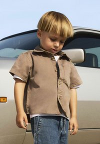 Chemise brune à manches courtes avec une poche avant, superposée sur un T-shirt blanc, associée à un jean en denim bleu. Design simple et décontracté.
