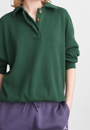 Kvinde iført en mørkegrøn tekstureret pullover med træknapper og lilla bukser med snøre, hænderne i lommerne, foran en ensfarvet baggrund.