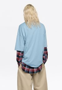 Camisa de manga corta azul claro superpuesta sobre una franela a cuadros roja y negra. Ajuste casual, estilo relajado, con pantalones color crema.