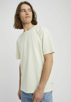 Jeune homme aux cheveux bruns mi-longs portant un t-shirt beige clair uni à manches courtes et un jean bleu, regardant vers sa gauche.