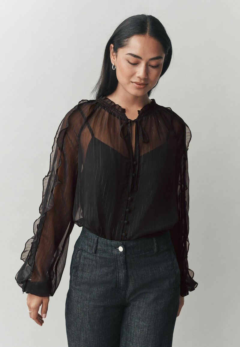 Blusa nera trasparente con polsini e colletto a volant, scollatura arricciata e dettagli con bottoni. Abbinata a pantaloni scuri e sartoriali.