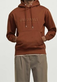 Sudadera marrón con bolsillo frontal, capucha con cordones y logo en relieve "ORIGINALS JACK & JONES". Combinada con pantalones beige.