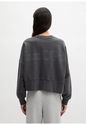 Femme aux longs cheveux bruns portant un sweat-shirt ample gris foncé avec le texte "Marc O'Polo" dans le dos et un pantalon gris clair, debout de dos.