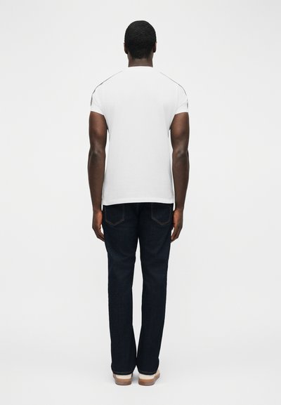 Άνδρας όρθιος, με πλάτη γυρισμένη, φοράει απλό λευκό κοντομάνικο πουκάμισο, σκούρα τζιν και ανοιχτόχρωμα παπούτσια πάνω σε λευκό φόντο.