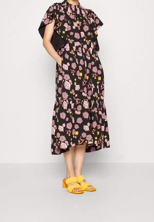 Vrouw draagt een zwarte midi-jurk met bloemenprint en gerimpelde mouwen en gele sandalen met open tenen en blokhakken, staat met haar handen in de zakken.