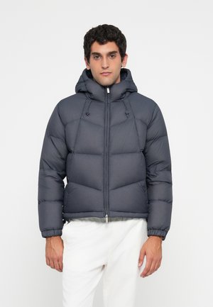 Homme portant une veste à capuche rembourrée grise foncée avec fermeture éclair et un pantalon blanc, se tenant devant un fond clair.
