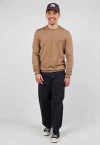 Pull en tricot marron clair avec un col rond, associé à un pantalon bleu marine foncé et des baskets noires. La tenue comprend une casquette de baseball sombre.