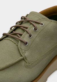 Ανοιχτό πράσινο σουέτ παπούτσι με στρογγυλή μύτη. Διαθέτει καφέ κορδόνια μέσα από ασημένια ροδέλες και διακριτικές ραφές. Τακούνι σε ταμπά απόχρωση με υφή στην επιφάνεια.