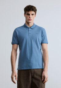 Gaiši zils īss piedurkņu polo krekls no kokvilnas, ar apkakli un trīs pogu priekšpusi, ar smalku logotipa izšuvumu uz krūtīm.