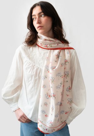 Écharpe en soie crème avec motifs floraux et théière, bordée de rouge. Texture légère et douce, elle se drape élégamment autour du cou.