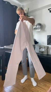 Homem com cabelo claro segura calças pálidas numa sala com chão de madeira, máquina de costura, capacete desportivo e uma cortina escura ao fundo.
