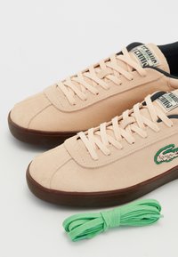 Baskets Lacoste en daim beige clair avec semelles foncées, lacets noués, logo sur le côté, et lacets verts supplémentaires à côté sur fond blanc.