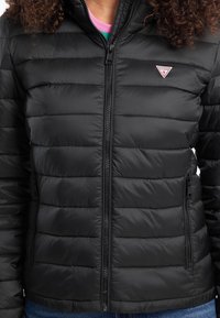 Svart puffjakke med horisontal quilted design, sentral glidelås og to glidelåslommer på sidene. Har en liten merkevarelogo på brystet.