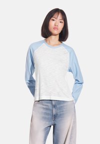 Koszulka raglan z długim rękawem o białym korpusie i jasnoniebieskich rękawach, z miękką fakturą i zaokrąglonym dołem. Luźny krój, minimalistyczny design.