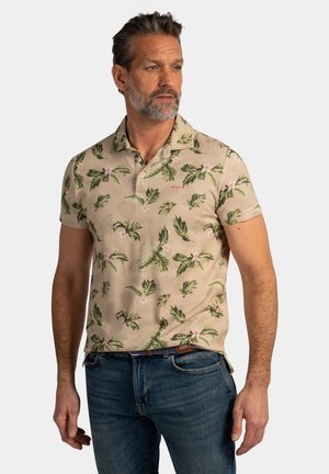 Man met grijze baard die een beige polo met korte mouwen draagt, bedrukt met groene bladeren en roze bloemen, staand tegen een effen lichte achtergrond.
