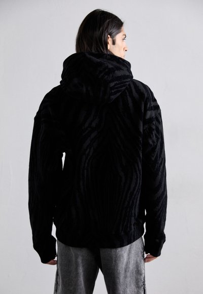 Hanorac negru cu un model texturat de dungi de tigru, având un glugă mare, mâneci lungi și o croială lejeră. Material moale cu un aspect plush.