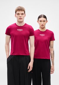 Vínové bavlněné trička s kulatým výstřihem a krátkými rukávy, s bílým logem "TOMMY JEANS" na hrudi. Kombinována s černými kalhotami.