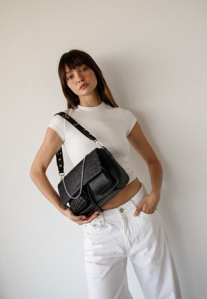 Jeune femme portant un t-shirt blanc court et un pantalon, tenant un sac à bandoulière en cuir noir texturé avec détail en chaîne, posée devant un mur uni.