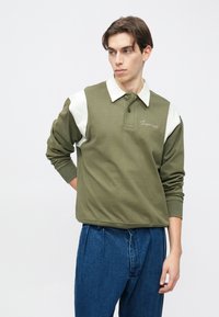 Polo de manga larga color verde oliva con un collar blanco y acentos en los hombros. Cuenta con cuello abotonado y texto bordado en el pecho.