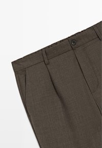 Gros plan sur un pantalon marron sur mesure avec fermeture à boutons, passants pour ceinture et poches latérales sur fond blanc.