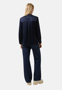 Marineblauwe blouse met lange mouwen, een satijnen pasvorm, afgeronde zoom en plooidetail aan de achterkant, gecombineerd met donker denim wijd uitlopende jeans en beige plateauzolen.
