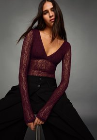 Top z bordovej čipky s dlhými rukávmi a hlbokým výstrihom do V, doplnený o čierne nohavice s vysokým pásom, veľkými gombíkmi a plisovaným dizajnom.