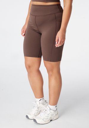 Vrouw met bruine high-waist fietsbroek met zijzak en witte sportschoenen en sokken, staand op een witte achtergrond.