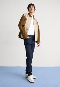 Beige and brown color-blocked jacket, white T-shirt, dark jeans, and light sneakers. Smooth fabric with a relaxed fit and button details.