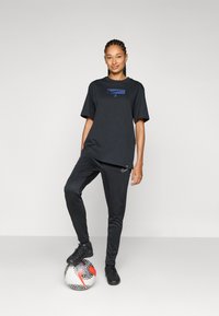 Black athletic t-shirt with a blue graphic, paired with black pants. The model stands on a patterned soccer ball, wearing black sneakers.