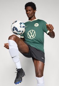 Zöld focimez világos gallérral és VW logóval, fekete nadrág, fehér zokni és fekete cipő. Játékos egy mintás focilabdán egyensúlyozik.