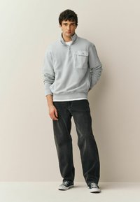 Lysegrå hættetrøje med lynlås og en lomme foran, kombineret med mørke jeans og sorte sneakers. Stoffet virker blødt og tekstureret.