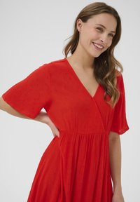 Robe rouge à manches courtes, décolleté en V, et tissu texturé avec des points en relief. Design fluide avec une taille froncée et une jupe ample.
