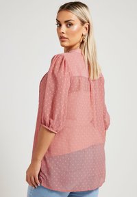 Blusa rosa trasparente con maniche a sbuffo, texture a pois e schiena parzialmente trasparente. Caratterizzata da un colletto rotondo e spacchi laterali.