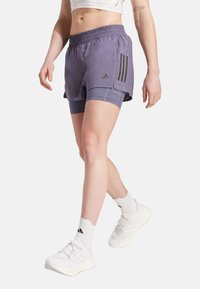 Lila idrottsshorts med ett elastiskt midjeband, med svarta tre-stripes-accenter och en lagerdesign, som bärs över grå kompressionsshorts.