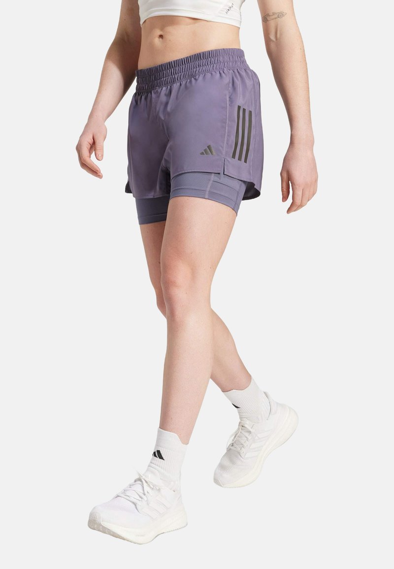 Lila idrottsshorts med ett elastiskt midjeband, med svarta tre-stripes-accenter och en lagerdesign, som bärs över grå kompressionsshorts.
