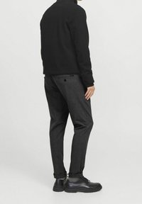 Maglione nero con colletto alto, abbinato a pantaloni grigi scuri con una texture sottile. Scarpe nere stringate completano l'outfit.