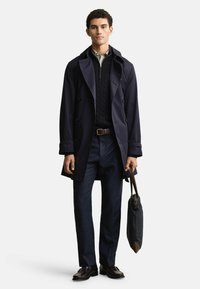 Navy trench coat z podwójnymi guzikami, nałożony na teksturowany czarny sweter. Ciemne spodnie i skórzane buty. Trzyma ciemną torbę.